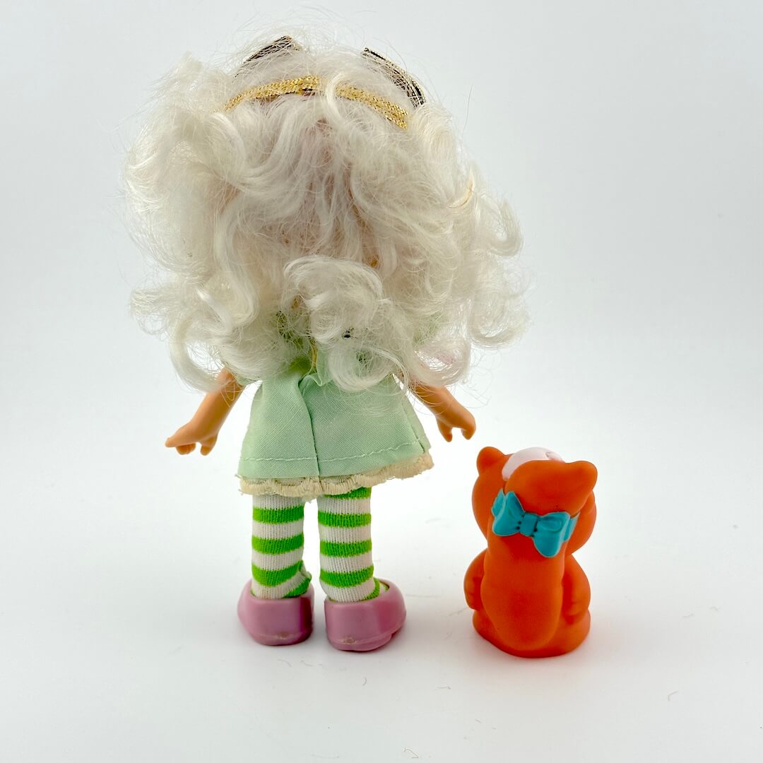 Toy doll with white hair and green outfit standing next to a small orange creature on a white background