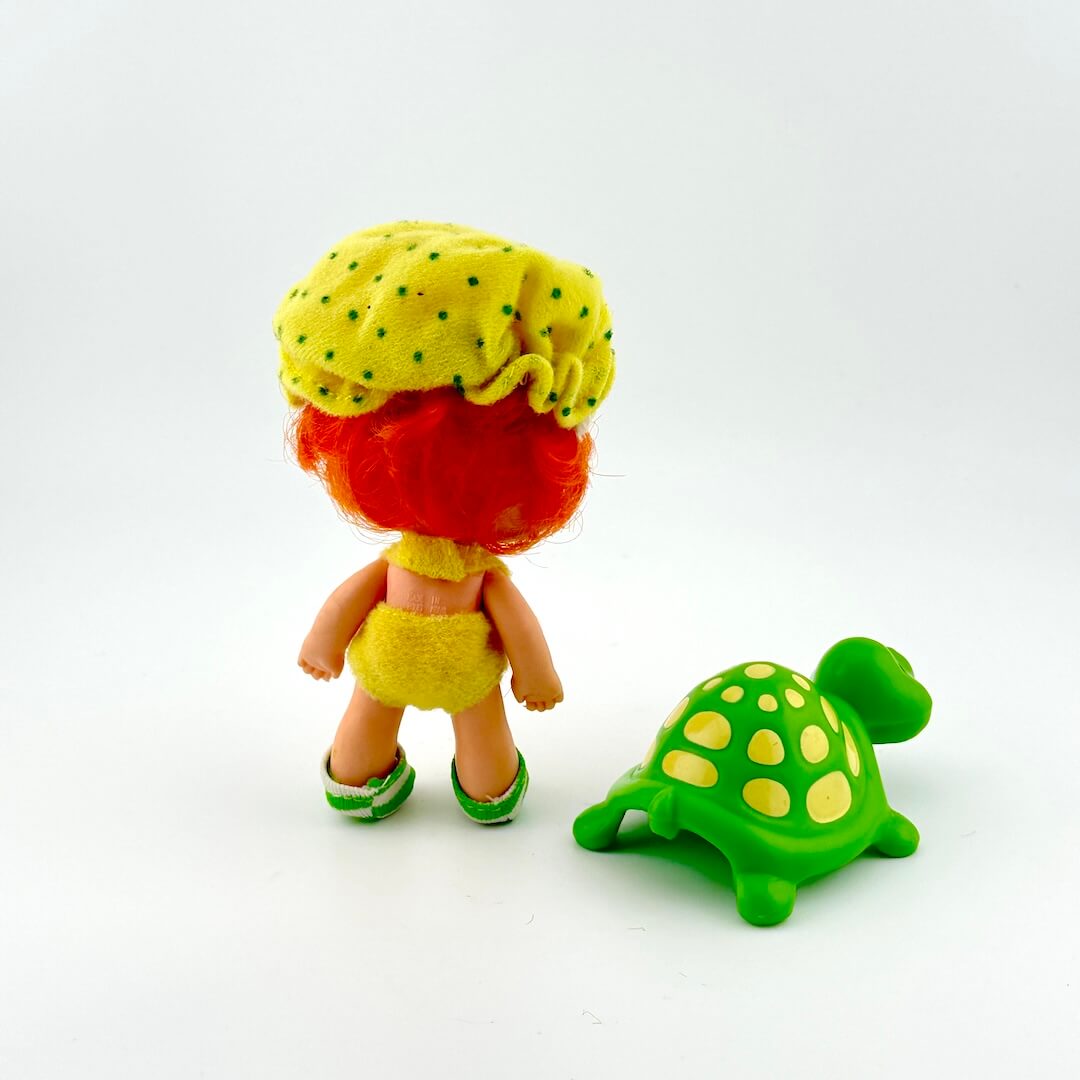Toy figure with red hair and yellow outfit standing next to a green turtle toy on a white background