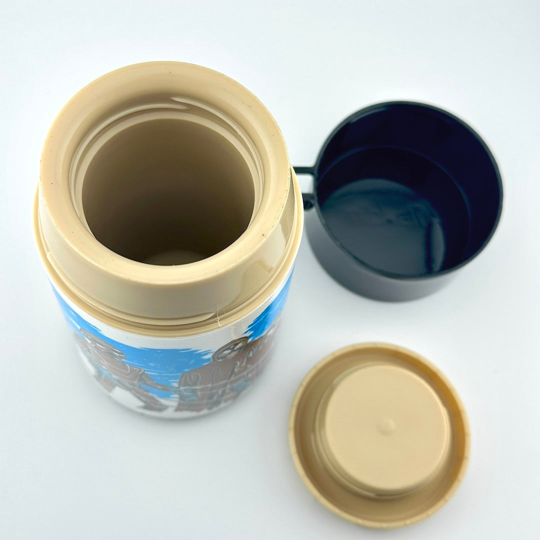 A thermos bottle with its lid off, showing a beige inner liner & a printed design, alongside a black lid & a separate beige lid.