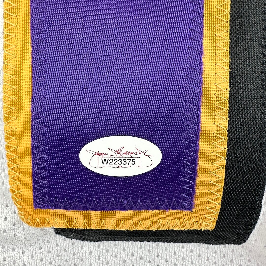 Close-up of a sports jersey with a purple number and yellow border and authentication sticker on a white jersey background