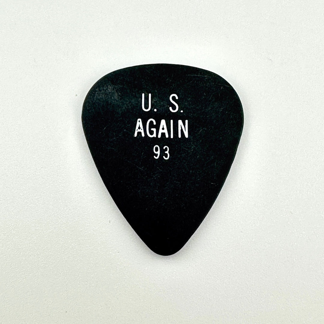 A black guitar pick with white printed text reading 'U. S. AGAIN 93' in the center, shown on a plain light background.