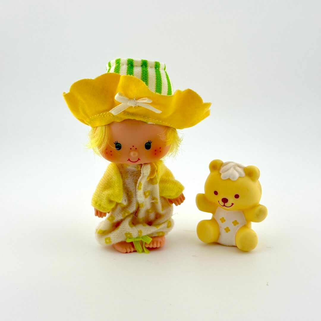 Toy figure of a girl with a yellow hat and dress, standing next to a toy bear on a white background