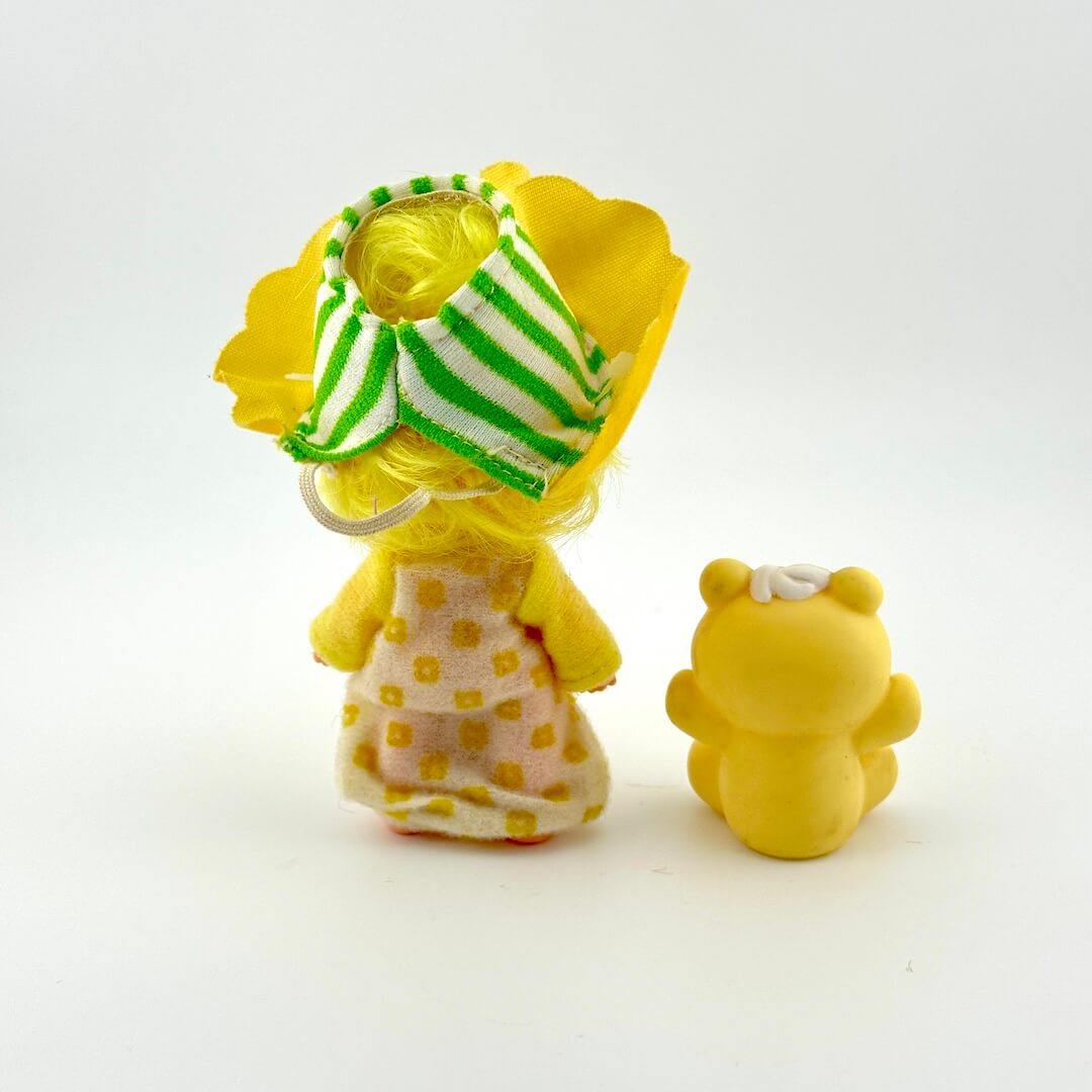 Two small figurines, one with a yellow dress and green and white striped hat, the other resembling a bear, on a white background.
