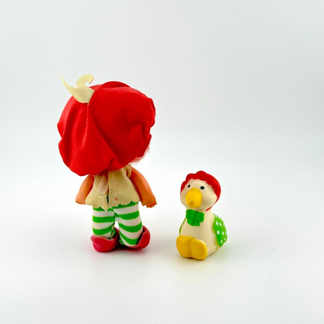 Two small toys, one with a red hat and green striped pants, the goose is smaller with a yellow beak and green outfit, on a white background.