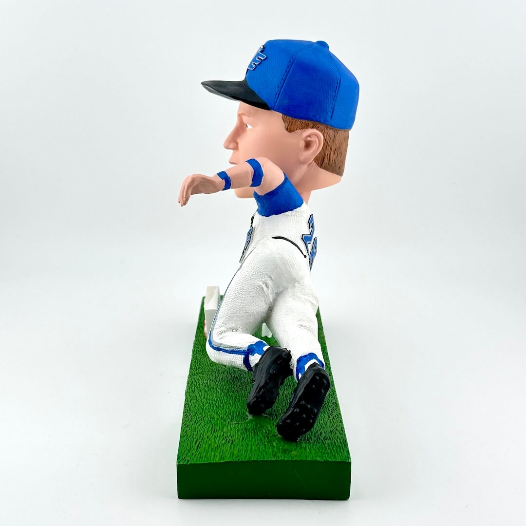 Bobblehead figure of a person in a blue cap and white outfit on a green base against a light gray background