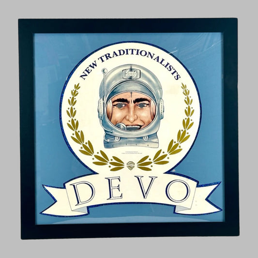 Black frame, blue matting, white circle with gold laurel ringed astronaut at center above "DEVO" in navy text on white banner