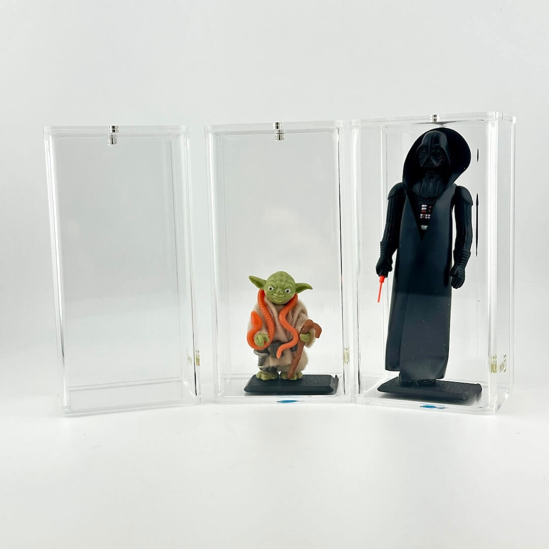 Two action figures, one green and one black, in a clear display case next to one empty case on a white background.
