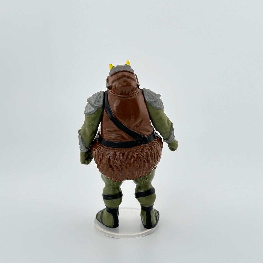 3-inch green hog-like alien with small horns wearing brown leather looking romper, silver armor on shoulders, black sandals.
