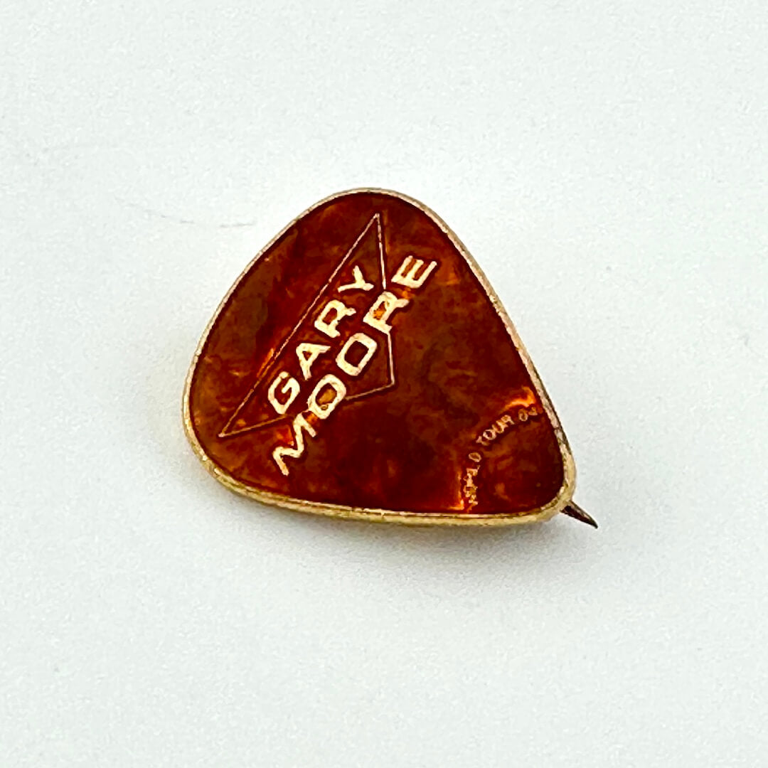 Brown enamel pin shaped like guitar pick with gold text, "Gary Moore, World Tour 84"