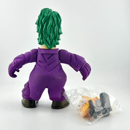 Toy figure of a character with green hair and purple outfit, standing next to a clear bag with additional accessories on a white background.