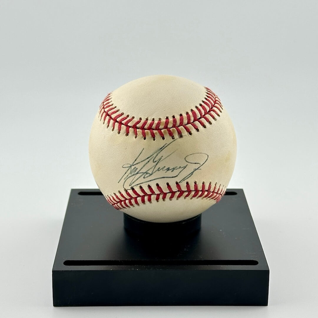 Autographed baseball in narrow space between seams sitting on top of black. base with two grooves for cover.
