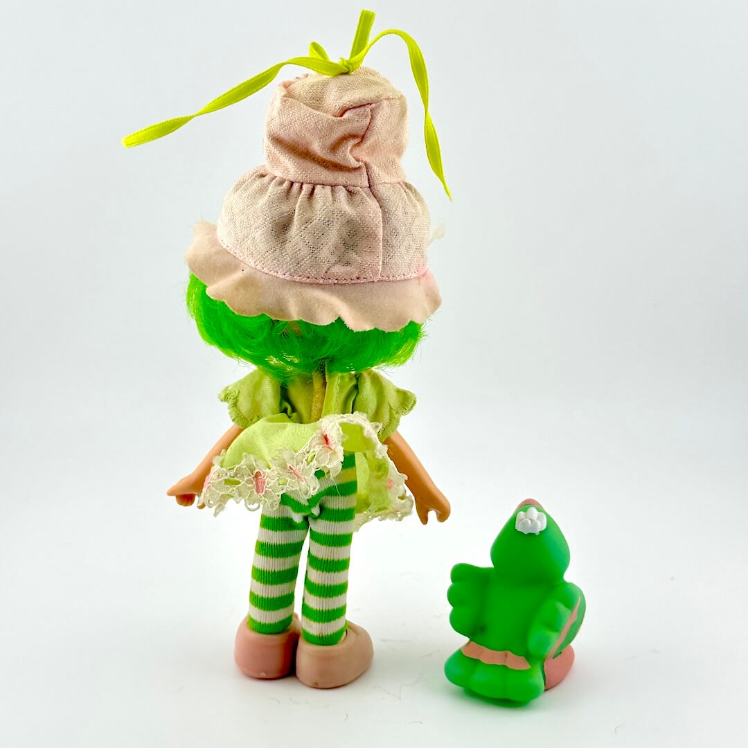 Toy doll in green dress and pink hat with green bird by side against white background.