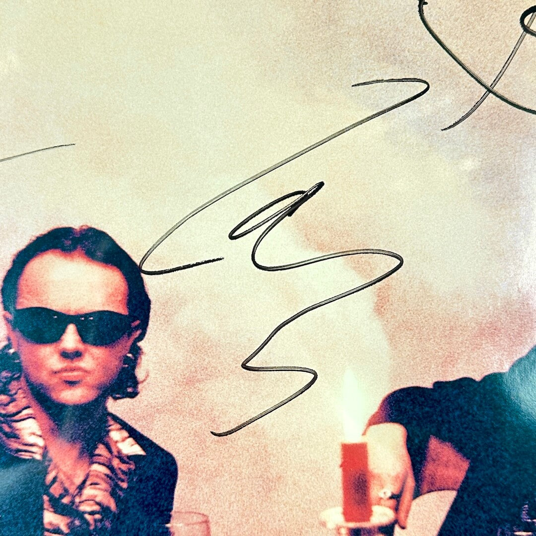 Person wearing sunglasses with a stylized signature above, sitting at a table with a drink.