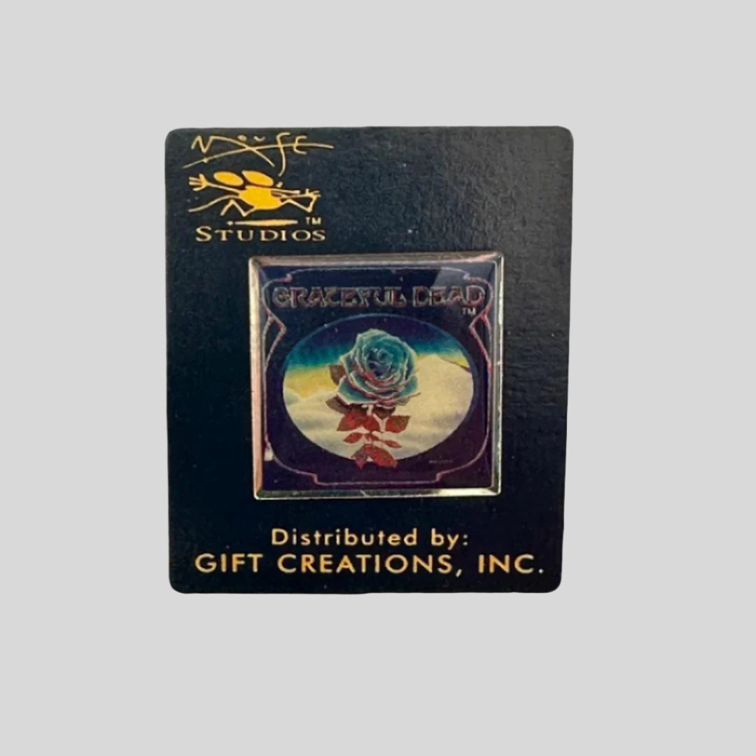 Hard enamel pin with blue rose with red leaves and stem in front of snow dunes with dark blue border.