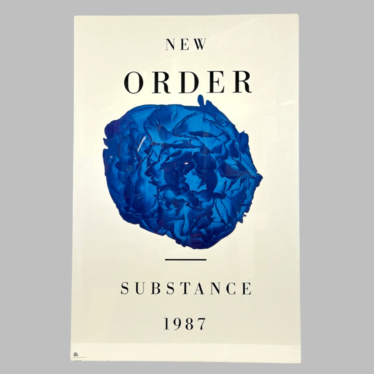 White poster with black text "New Order" at top above central blue flower, "Substance, 1987" below.