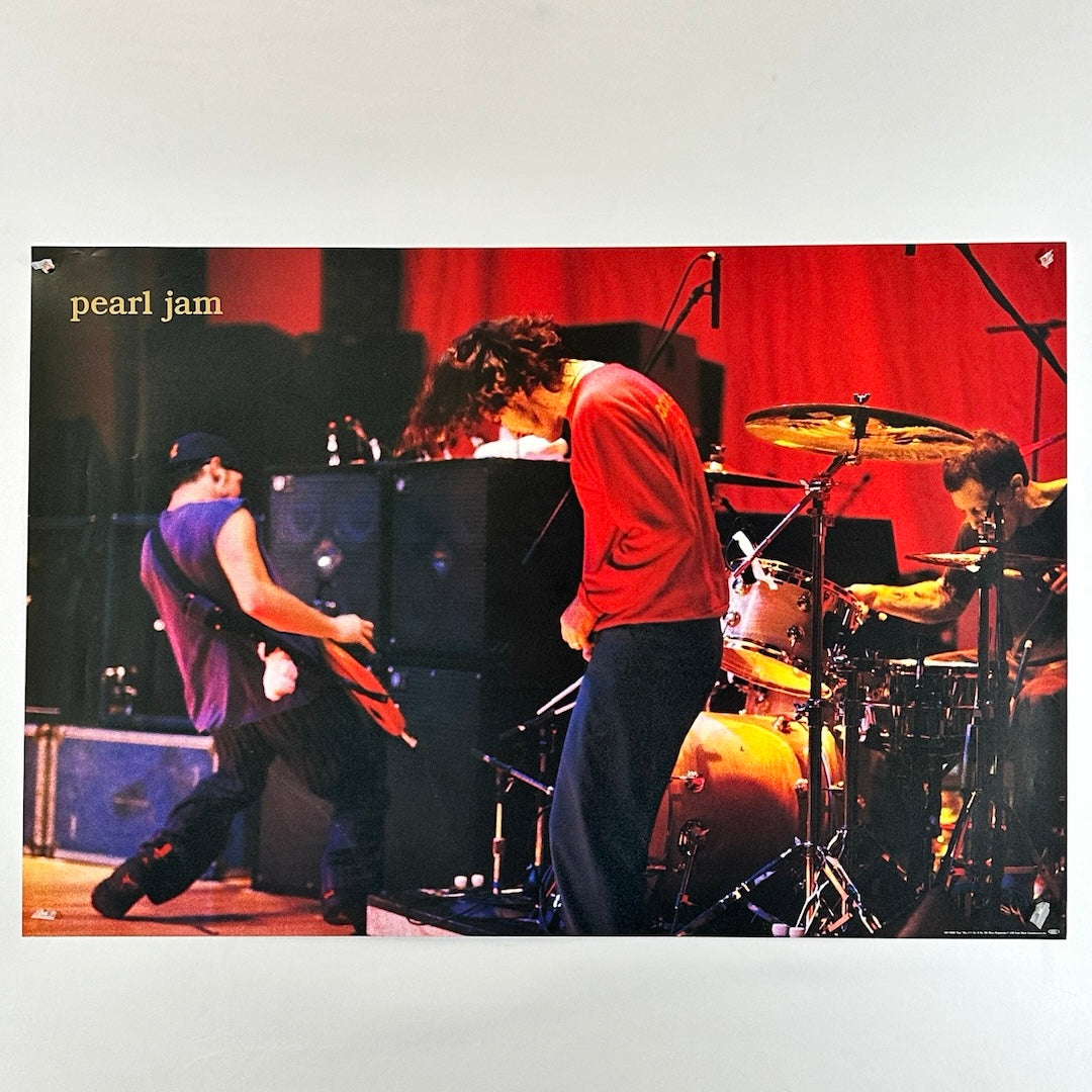 Poster of Pearl Jam playing music together on stage with red backdrop. Guitarists are headbanging, & drummer also visible.