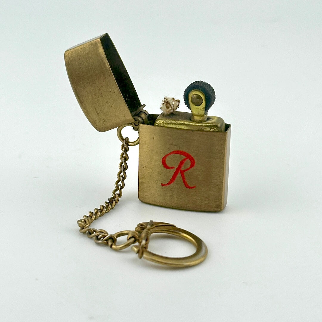 A miniature brass pocket lighter keychain with an open lid, a small chain, and a key ring, featuring a red letter 'R' on the front.