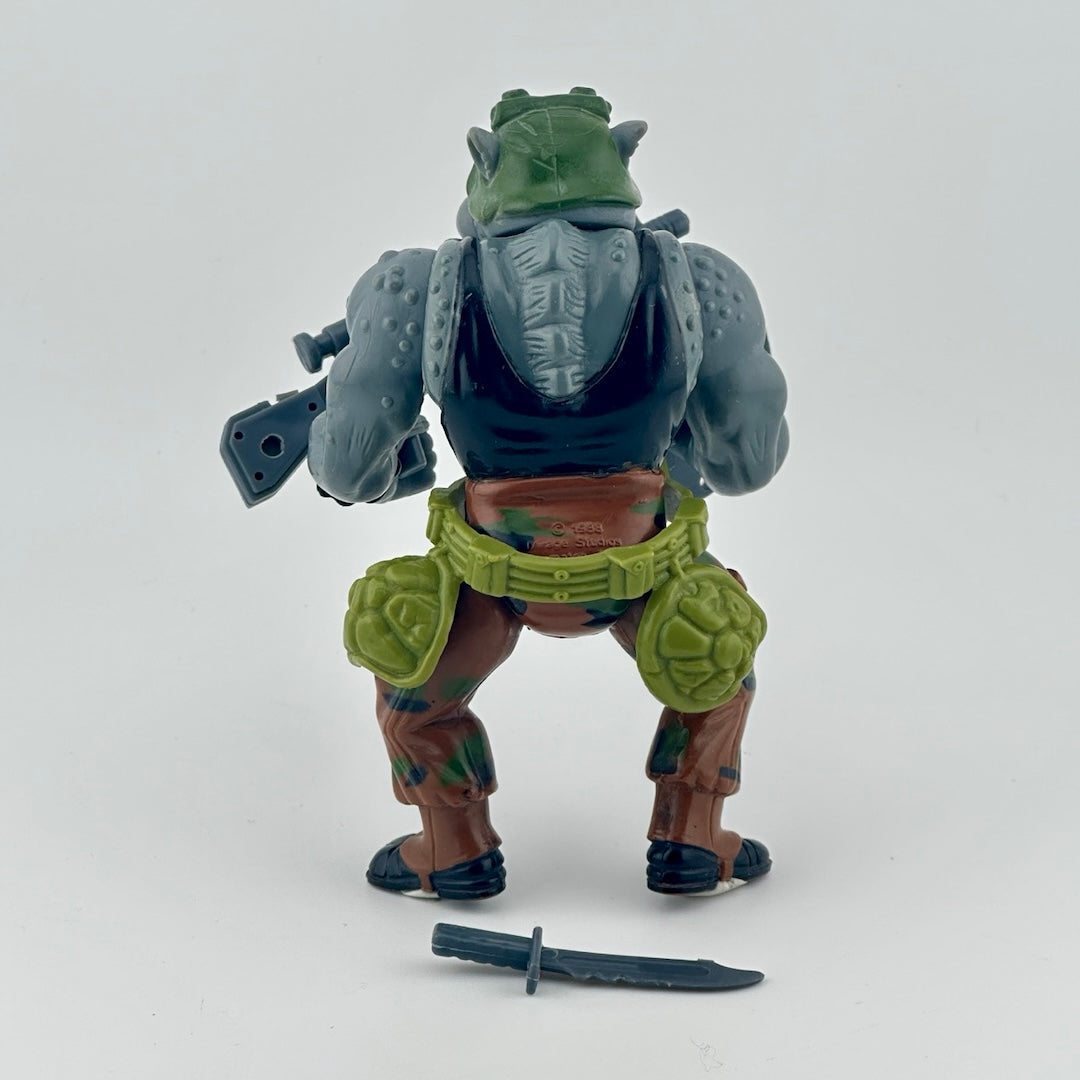 Anthropomorphized Rhino with in brown outfit, green belt with turtle shells, grey trash can lid shield, grey gun, and grey knife. Back.