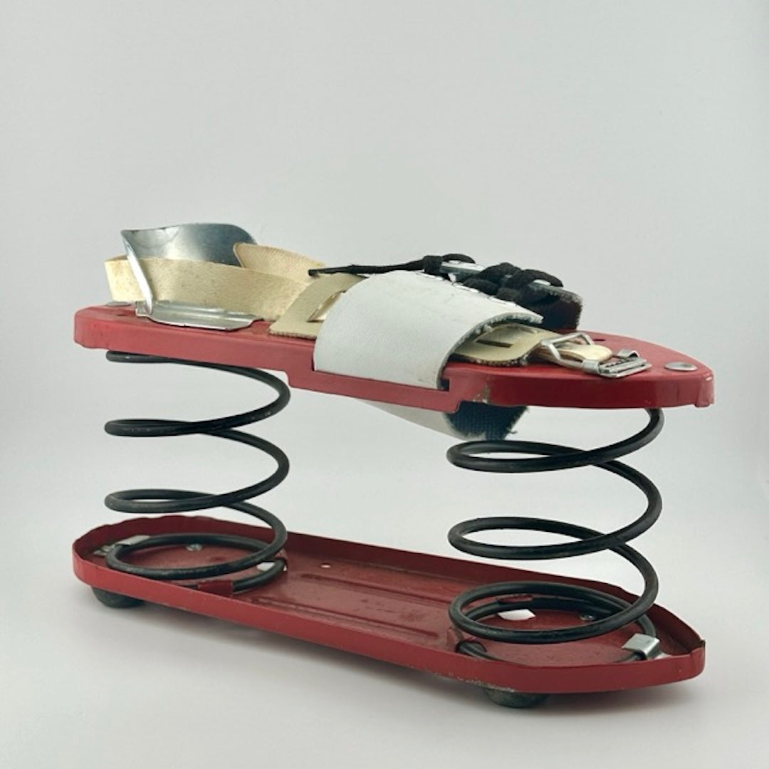 Oval red metal platforms separated by two large springs. White leather straps across top with laces, metal heel piece.