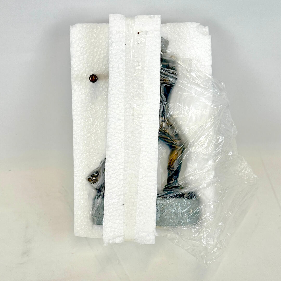 A small dark figurine wrapped in clear plastic and resting on white foam inserts.