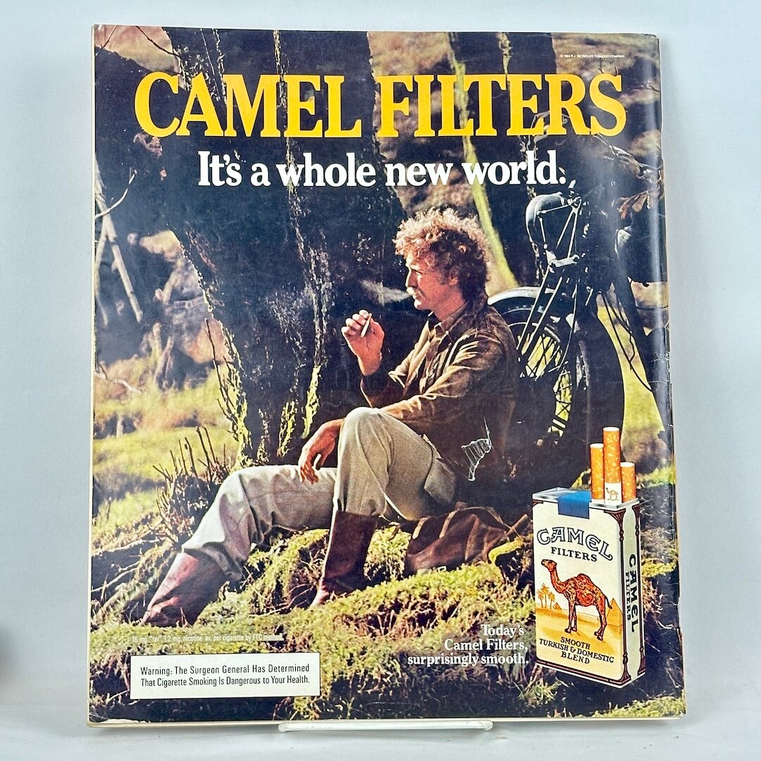 Back cover of magazine with full page ad for Camel cigarettes featuring smoking man, leaning against a tree by motorcycle. 