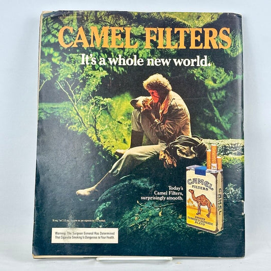 Back cover of magazine with full page ad for Camel cigarettes with image of man smoking on mossy tree branch. 