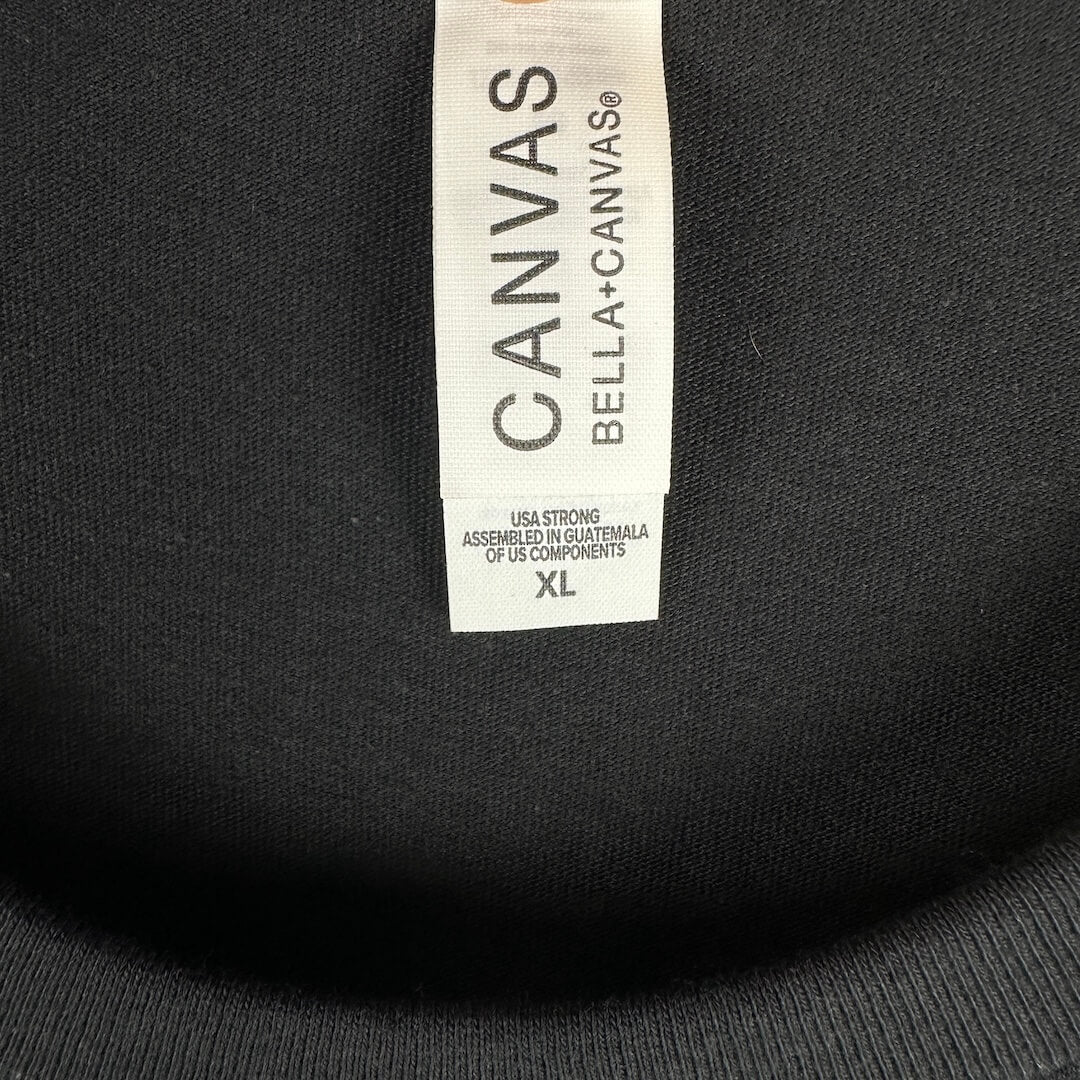 Close-up of a black garment's inner neck label reading CANVAS BELLA+CANVAS, with size XL printed on the white tag.