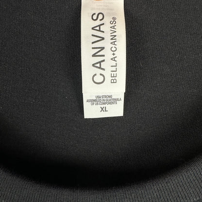 Close-up of a black garment's inner neck label reading CANVAS BELLA+CANVAS, with size XL printed on the white tag.