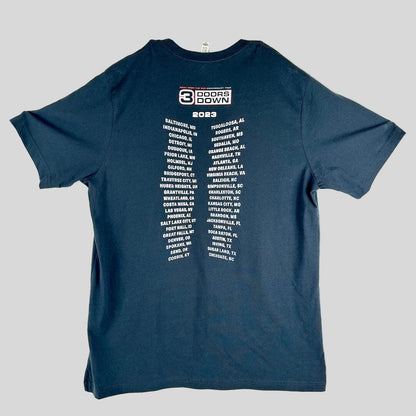 A black t-shirt laid flat, featuring a white graphic print of the 2023 '3 Doors Down' tour city list on the back.