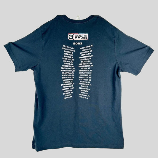 A black t-shirt laid flat, featuring a white graphic print of the 2023 '3 Doors Down' tour city list on the back.