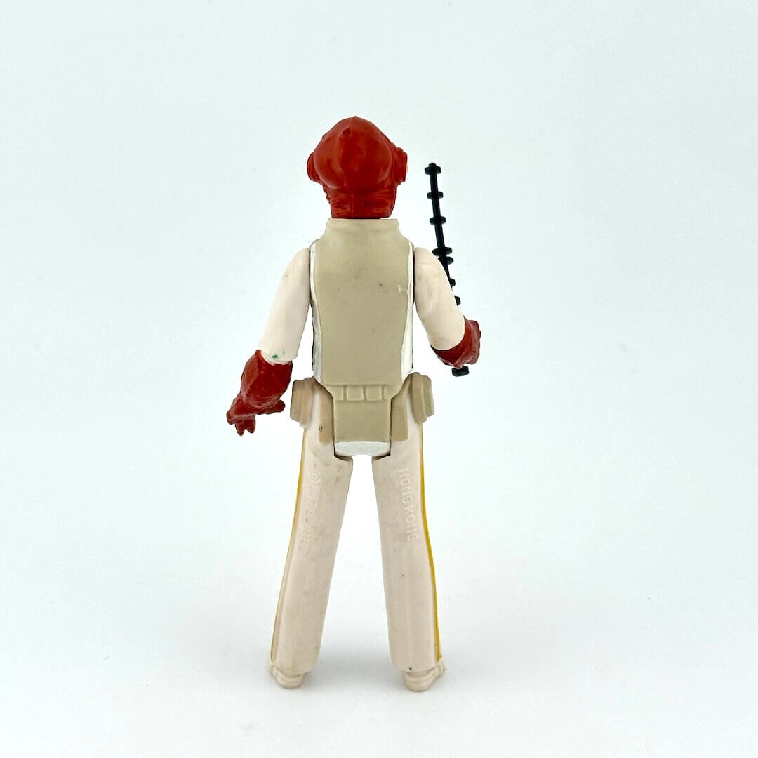 Action figure with red head and beige outfit holding a black and black weapon on a light gray background