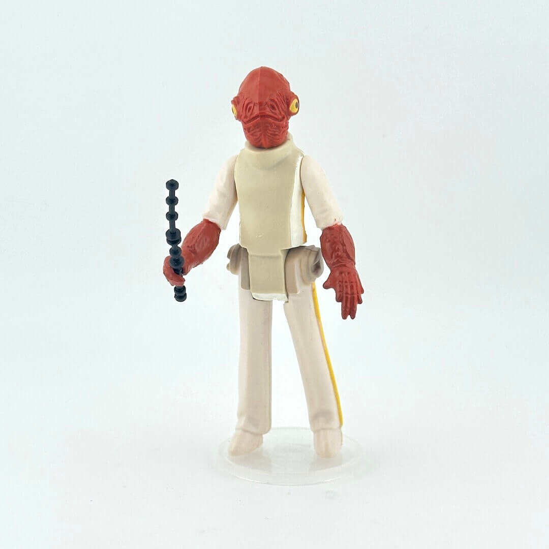 Action figure with red head and beige outfit holding a black and black weapon on a light gray background