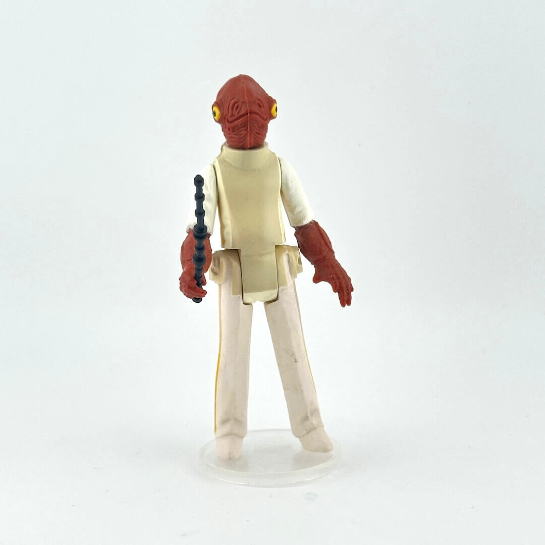Action figure with red head and beige outfit holding a black and black weapon on a light gray background