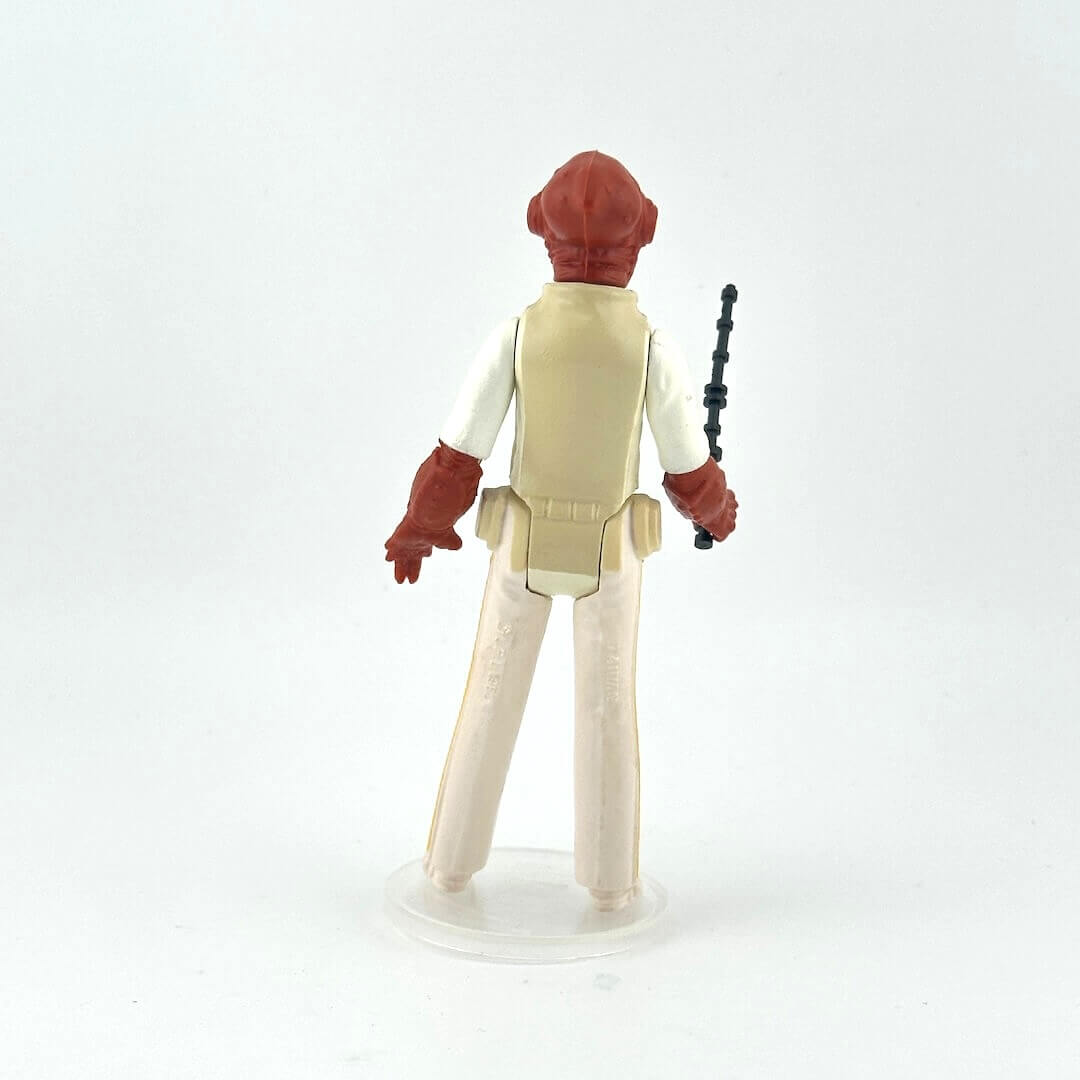 Action figure with red head and beige outfit holding a black and black weapon on a light gray background