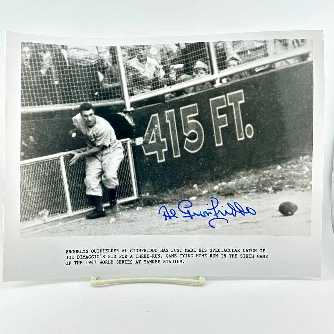 Black and white photograph of vintage baseball player mid-catch beside blue signature against white background. 