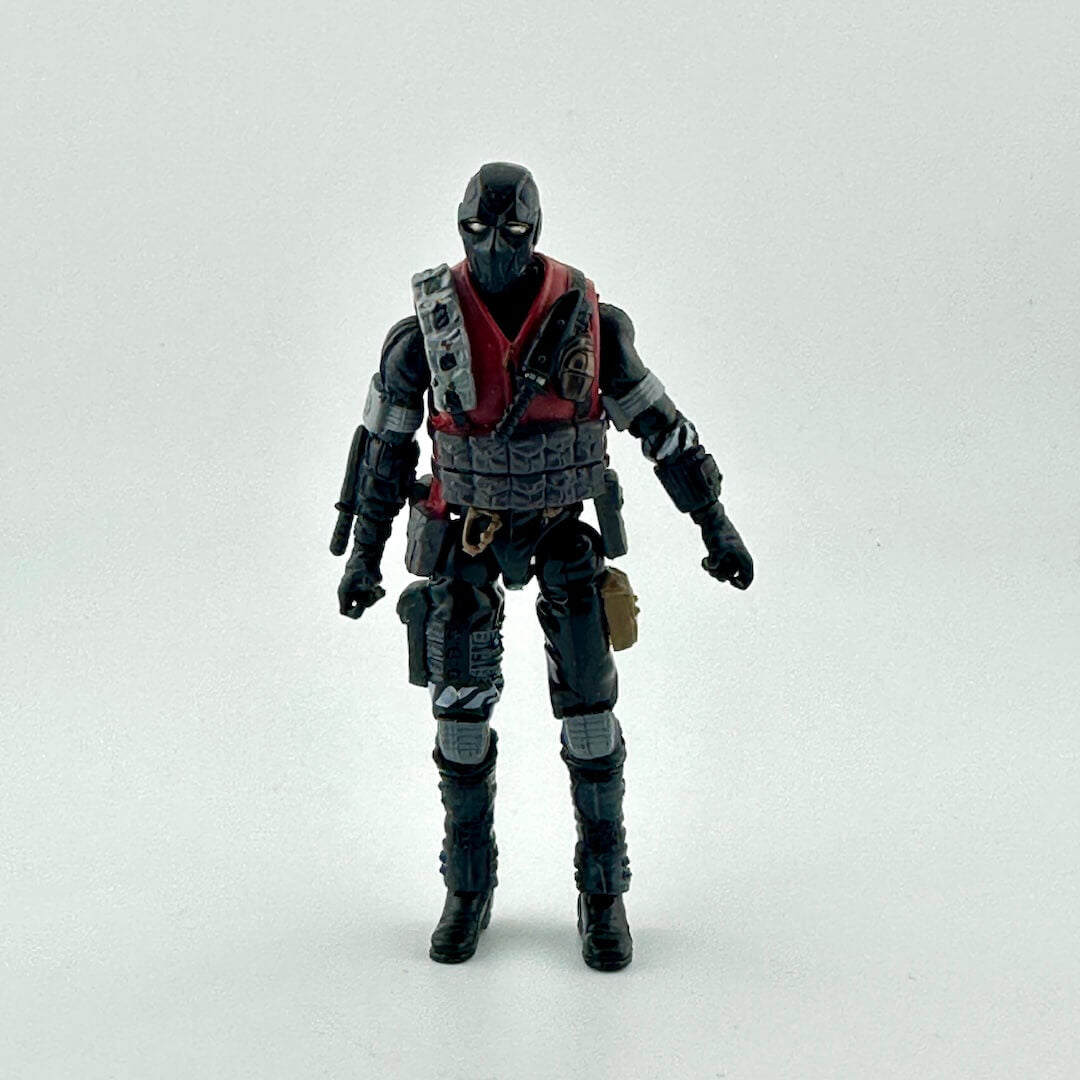 An action figure in all black with red accents wearing a black mask, standing in front of white background. 