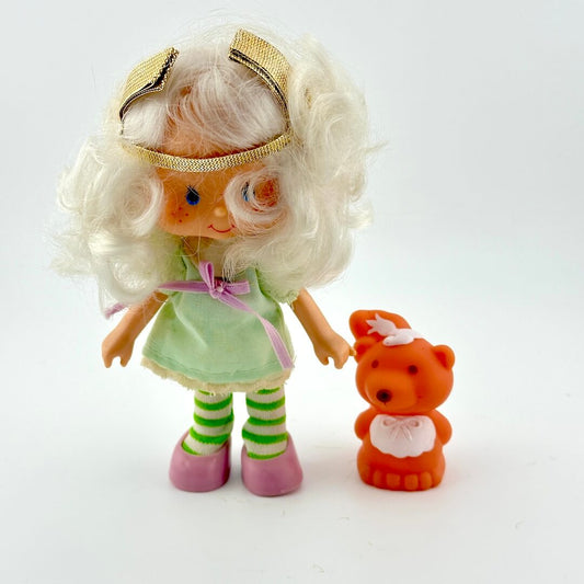 Toy figure with blonde hair, green dress, and striped socks holding a small orange bear toy on a white background.