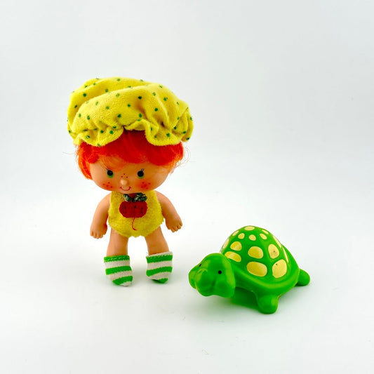 Toy doll in yellow onesie and green striped socks, standing by toy turtle. 