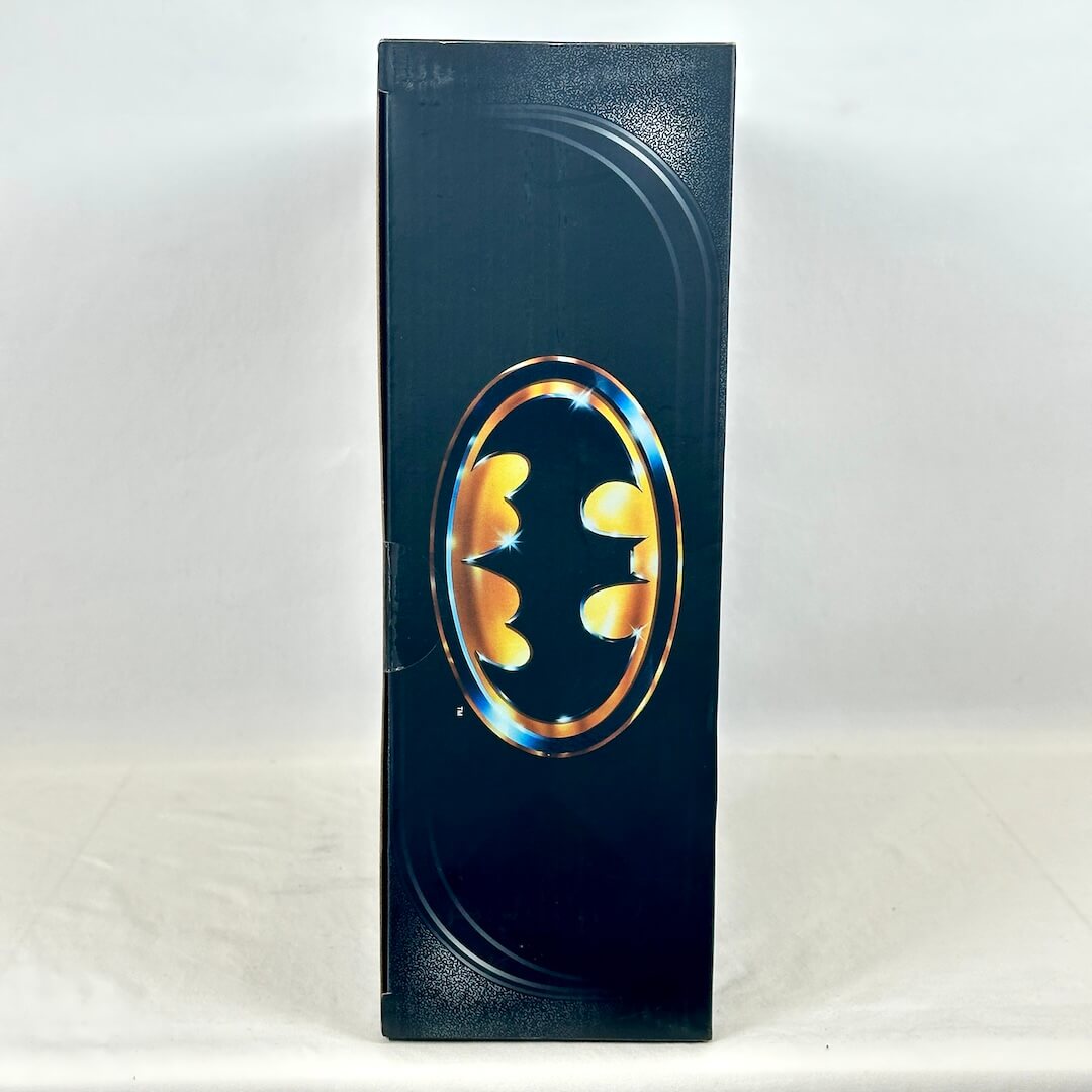 A tall, narrow black product box featuring a large metallic gold and blue Batman logo emblem on the front.