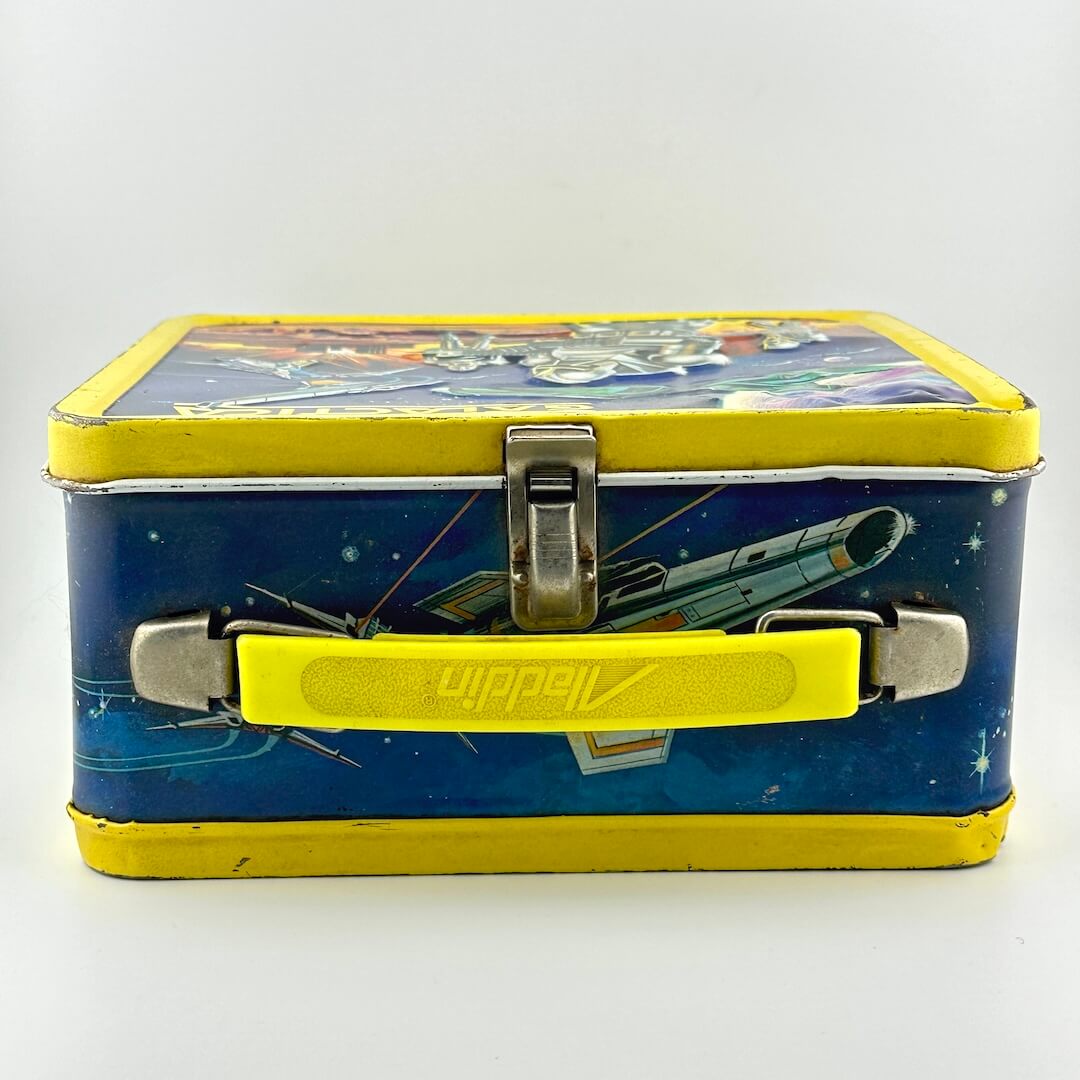 Top view of metal lunchbox with yellow plastic handle, yellow borders, blue background with spaceship illustration. 