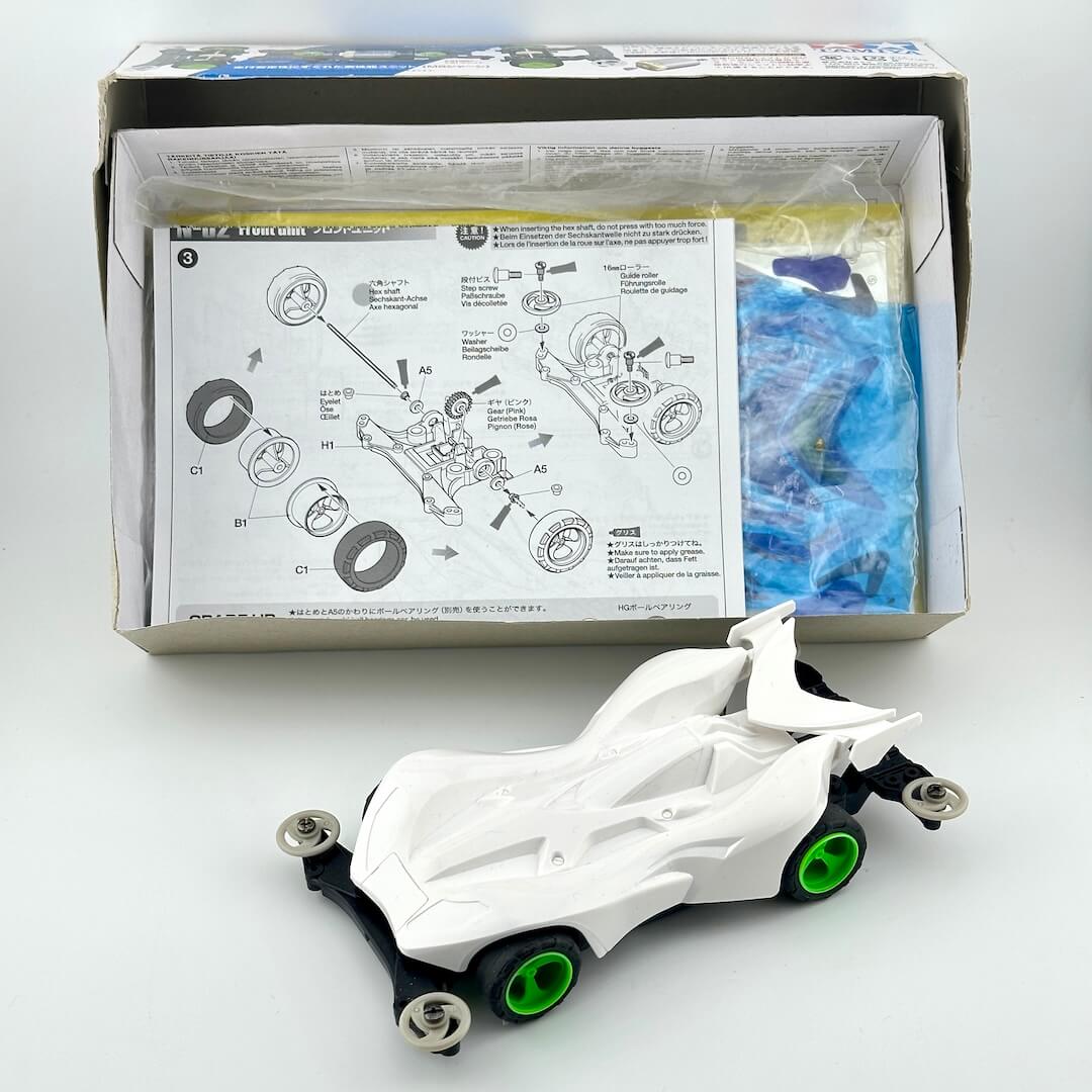 A white toy race car with green wheels displayed in front of its open packaging box containing an instruction manual and parts bag.