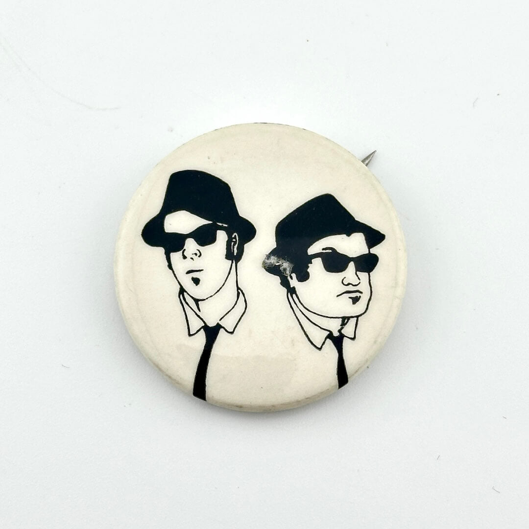 Button badge with illustration of two people wearing hats and sunglasses on a white background