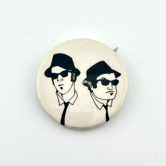 Button badge with illustration of two people wearing hats and sunglasses on a white background