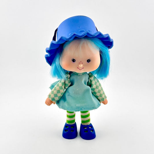 Toy doll with blue hat and hair in blue dress against white background.