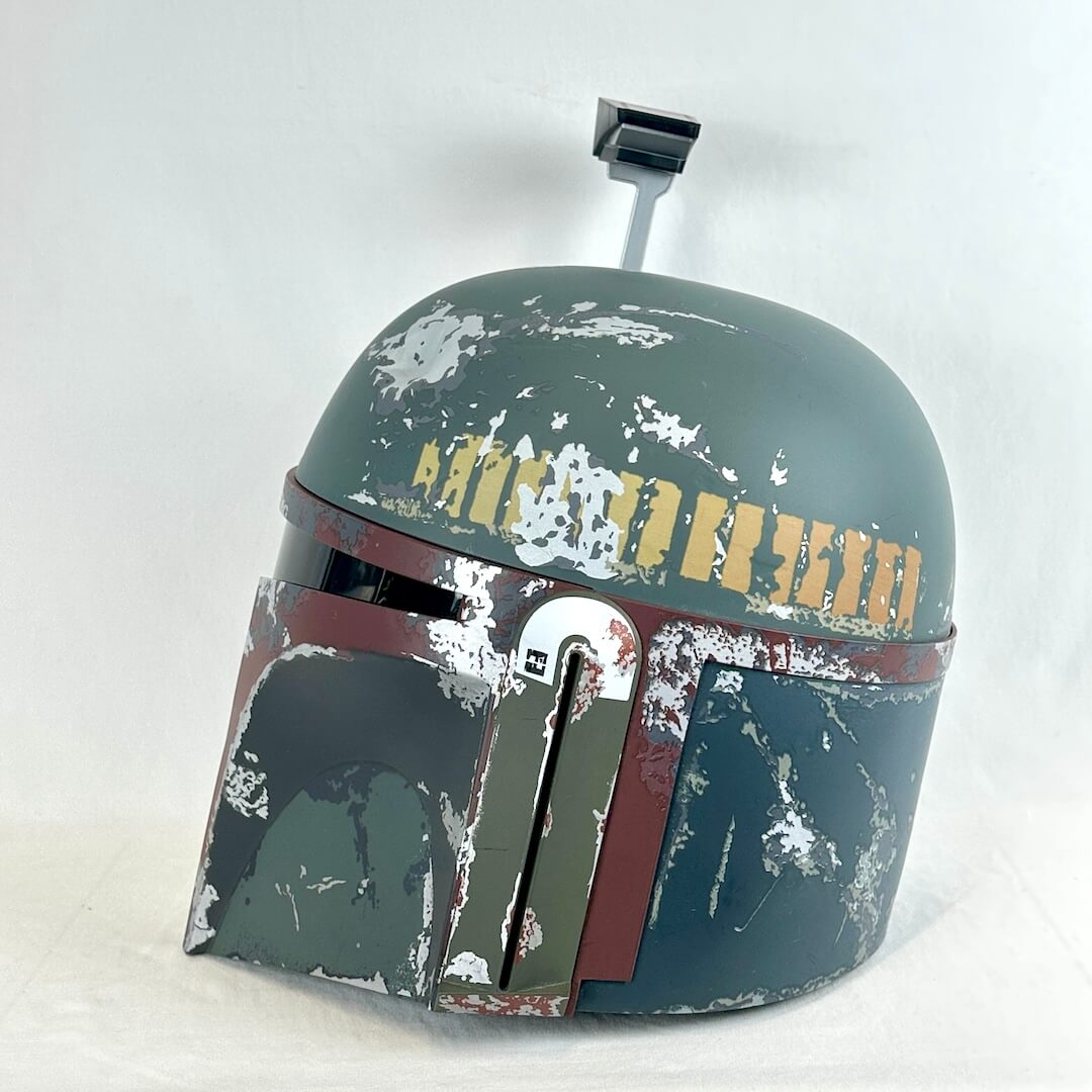 A Boba Fett style sci-fi helmet prop with a distressed green-gray finish, gold lettering, and weathered red and white paint details.