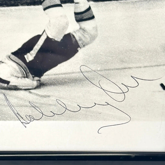 A black and white signed photograph of a hockey player on the ice, featuring a large cursive autograph across the lower portion.