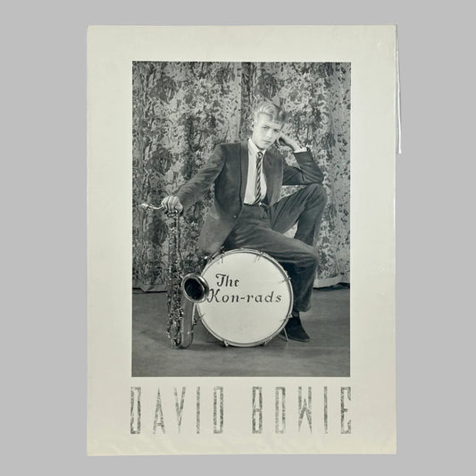 Black and white photo of young Bowie in suit, seated on a drum, leaning on a saxophone. Name along bottom. 