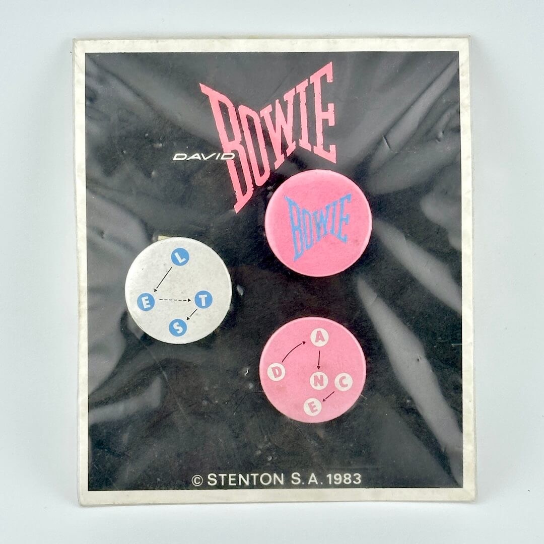 Black card with David Bowie branding with three circular pins in white and pink color scheme. 