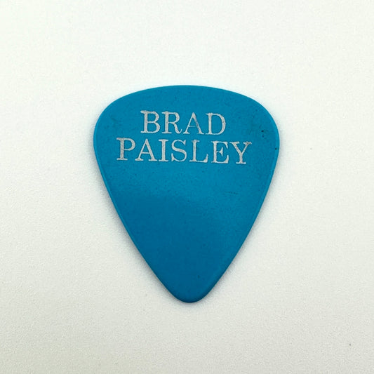 Blue guitar pick with silver text, "Brad Paisley" photographed against white background. 