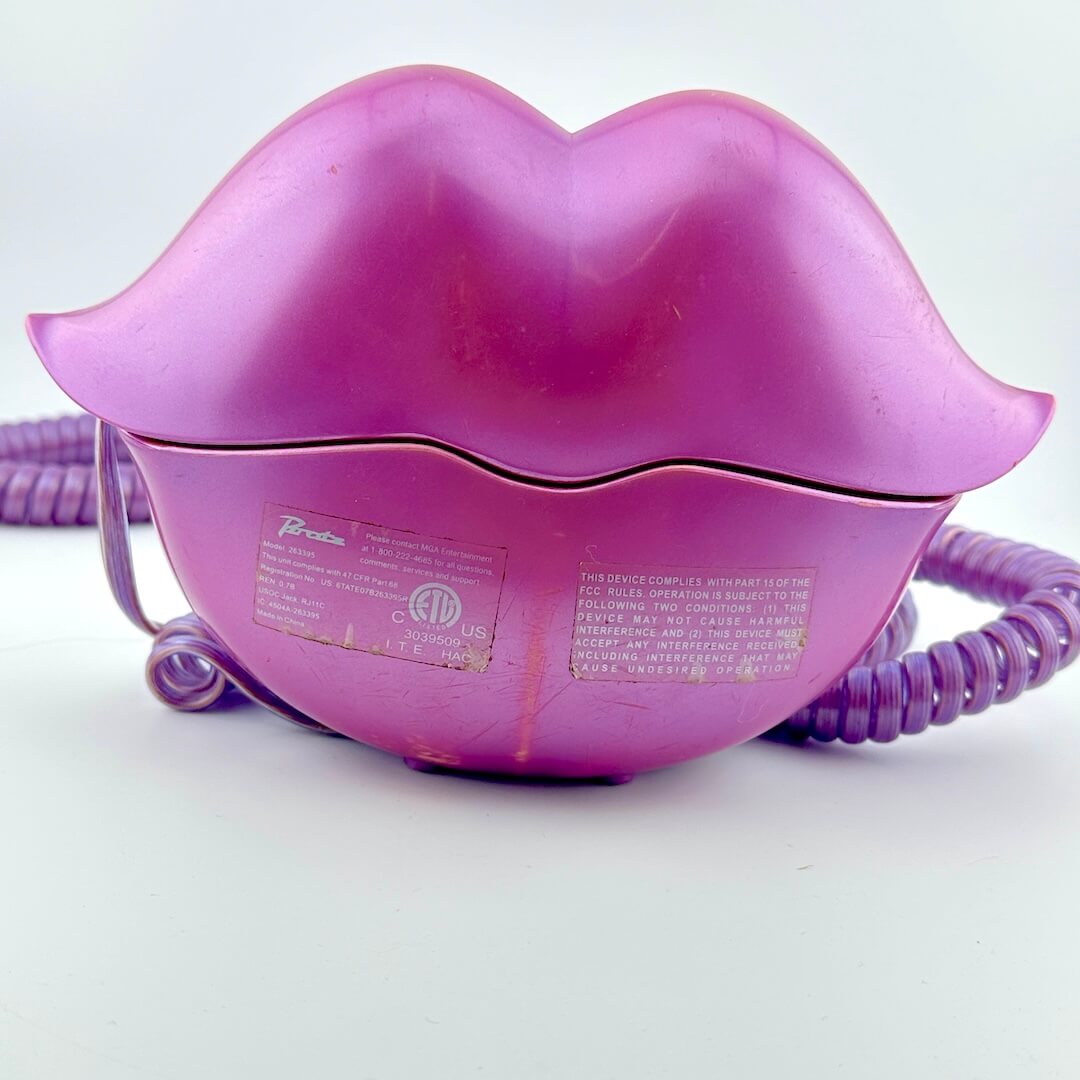 A purple plastic lip-shaped phone with purple coiled cord behind it, photographed on light background. 